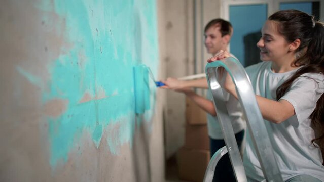 Guy and girl moved to new building, they doing repairs in their new apartment. Painters working, painting walls together with roller. Water primer or blue paint. Happy married couple. Home decoration.