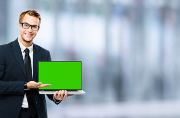 Business man in glasses spectacles, black suit show laptop with empty mockup green chroma key screen, on blurred office background. It expert,  technician repair service, tech support maintenance