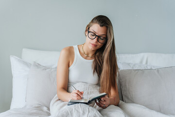 Young brunette caucasian woman in white t-shirt sits on bed writes in diary planning day, agenda. American student girl in glasses makes note, awakes home at bedroom. Daily routine, women.