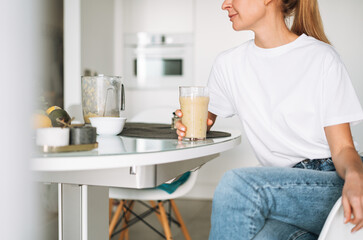 Young slim woman in white t-shirt and blue jeans drinking fruit smoothie healthy food in kitchen at...
