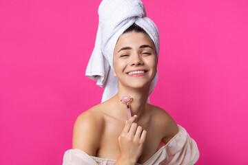 A beautiful cheerful attractive girl with a towel on her head holds a cosmetic roller near her face against a pink background.