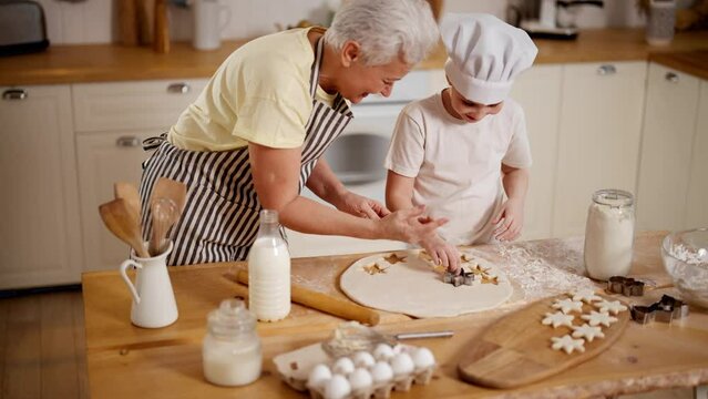 Granny and granddaughter pre-teen girl cooks homemade cookies in kitchen at home together. Family recipe, sweets and bakery, preparing fresh food. They squeeze out cookies star shape from dough.