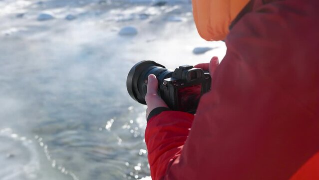 Detail of a man with a red jacket and camera exhaling and photographing small river on a winter sunny day.