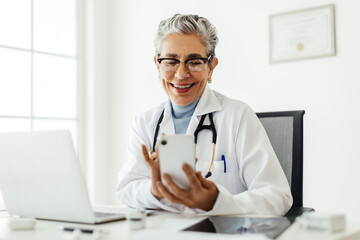The online doctor: Happy female physician video calling a patient on a smartphone - obrazy, fototapety, plakaty