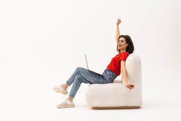 Euphoric young woman celebrating success with laptop, raising arms up, sitting in armchair on white...