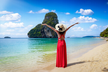 A happy tourist woman in a red dress stands at the beautiful Phra Nang beach at Krabi, Thailand, during her tropical holidays