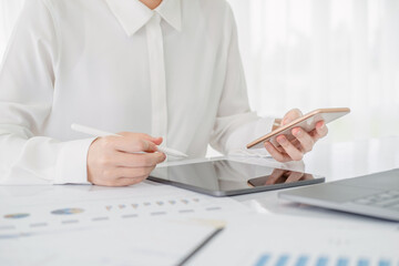 Asian businesswoman in white formal clothes writing note in tablet and checking smart phone. Business woman busy working with documents and tablet  in workstation. close up clean image.