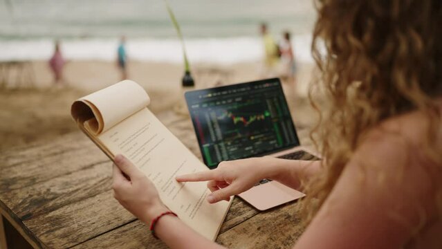 Female freelancer with laptop sitting in outdoor tropical cafe reading menu. Young adult woman working remotely at resort checks the menu choosing food. Female crypto trader working from resort