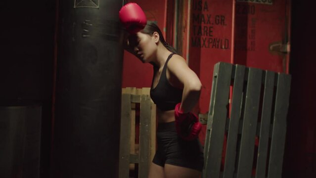 Tired breathless athletic fitness lovely Asian woman in boxing gloves taking a break, leaning on punching bag, feeling exhausted while improving boxing techniques and skills in dark garage gym.