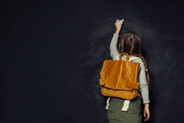 Child girl with backpack standing with her back and writing on school chalkboard. Back to school, education, student, pupil