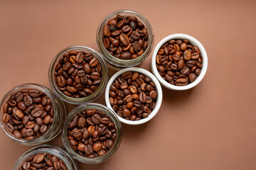 Aromatic roasted coffee beans in small glass jars and in white plates from above in beautiful minimalist composition on brown background. 