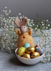 Crochet bunny and bowl of Easter sweets on a table. Colorful still life with chocolate eggs, flowers and toy. Easter celebration concept. 