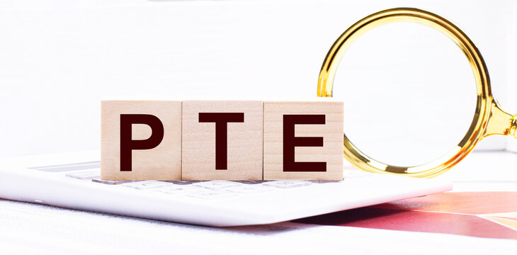 Three wooden cubes with the text PTE Pearson Test of English stand on a white calculator near a magnifying glass. Business concept