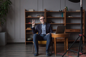 Portrait of mature serious businessman in suit and glasses is sitting in the office and showing peace sign