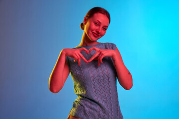 Portrait of adorable young girl with cute face showing crossed fingers in gesture of heart over blue background in neon light