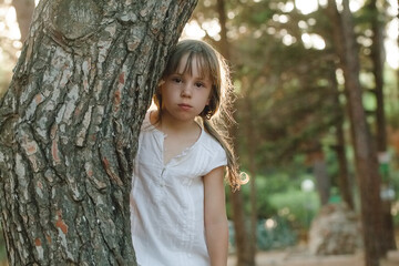Cute child girl in white clothes standing near tree in the park