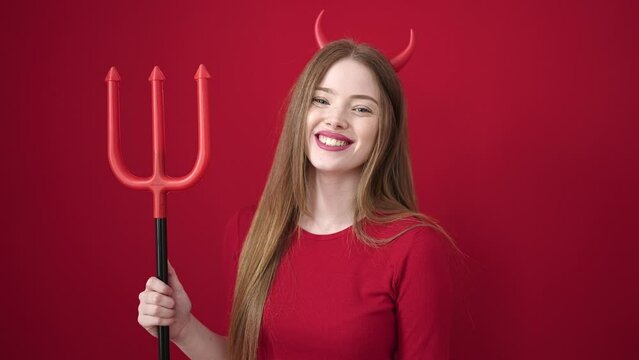 Young blonde woman wearing devil costume holding trident over isolated red background