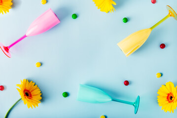 Top view background with bright plastic cocktail glasses, multicolor candy drops and yellow gerbera flowers on the light blue background. Summer party mood. Flatlay, copy space.