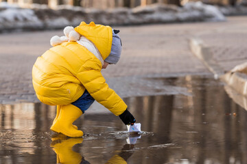 A small child in yellow rubber boots and a jacket runs through puddles, has fun, plays and launches paper boats. Spring break photo. It's springtime.