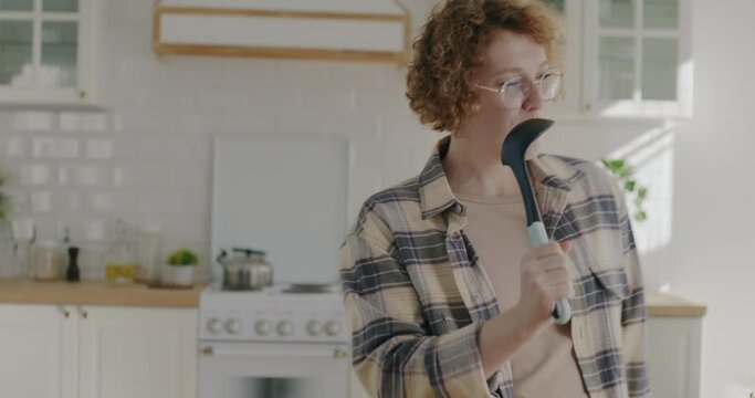 Portrait of playful young lady dancing and singing in spoon having fun in kitchen at home. Entertainment and modern lifestyle concept.
