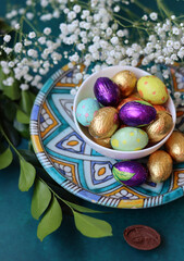 Foil wrapped Easter chocolate eggs in a glass bowl. Easter celebration concept. Colorful still life with sweets. Textured background with copy space. Easter greeting card. 