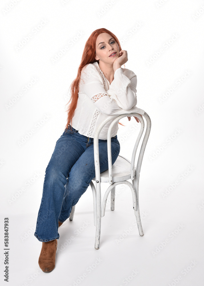 Sticker full length portrait of beautiful woman model with long red hair, wearing casual outfit white blouse  top and denim jeans, isolated on white studio background. Relaxed seated pose, sitting on chair wi