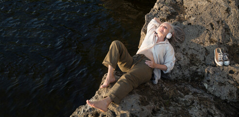 Summer lifestyle portrait senior woman  with gray hair in headphones relaxes on the seashore. spends time in nature in summer. Audio healing. meditation. banner. copy space. greyhairdontcare