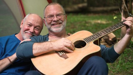 Closeup portrait of two gay men at campsite with tent and guitar turning and looking at camera.