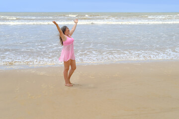 woman 50 years old with long hair in pink swimsuit stands barefoot on sandy seashore, enjoys sea breeze, takes sunbaths, blue waves, concept travel, vacation in tropical, summer sun protection