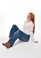 full length portrait of beautiful woman model with long red hair, wearing casual outfit white blouse  top and denim jeans, isolated on white studio background. Relaxed seated pose, sitting on floor wi