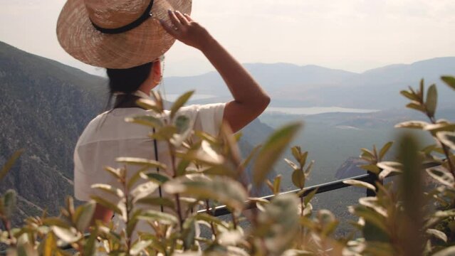 Young woman looking at Delphi landscape with mountains from high viewpoint at sunset, Greece. Fashion white dress, sunglasses, large hat.