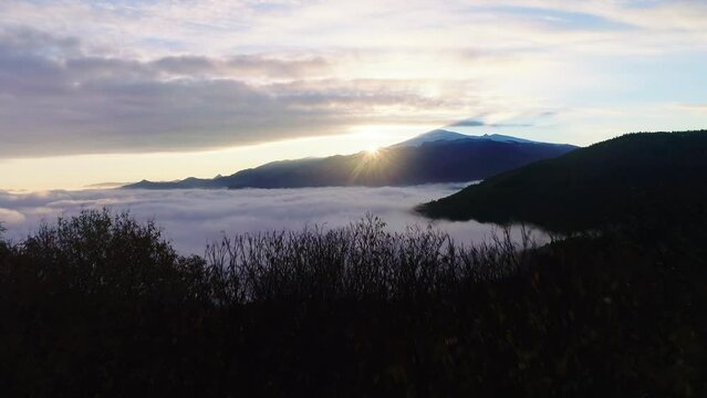 Flying over the trees to reveal the clouds and sunrise behind the mountain