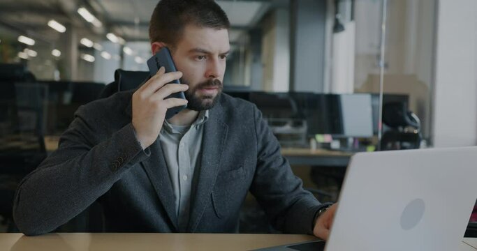 Serious businessman talking on mobile phone and yuping with laptop working in glass wall office. Business and modern technology concept.