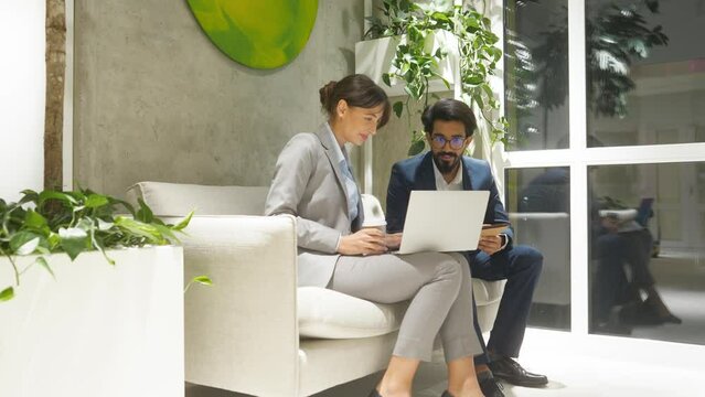 Portrait of two young attractive professional workers of modern company sitting on sofa in office terrace looking at laptop smiling. Stylish male Indian man and happy beautiful woman wearing suits.