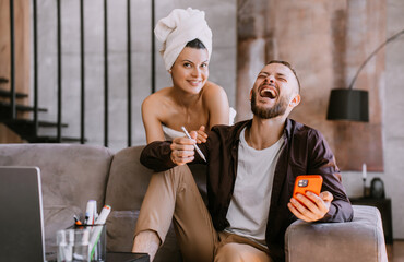 Young beardy caucasian man laughing out loud holds phone sits on couch home, pretty hispanic woman with towel wrapped around head looks at camera. Newlywed at honeymoon. Couple at hotel.