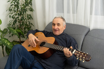 Senior man is playing guitar. Elderly man sitting on the sofa and playing guitar. Portrait of a gray-haired mature man in a sweater learning to play. Enjoying retirement life at home.