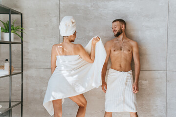 Back view on woman wrapped in towel  standing at bathroom showing body to husband being in playful mood. Newlyweds at honeymoon. Cheerful caucasian guy looks at wife after spa procedure.