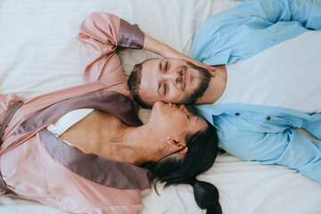 Top view on young caucasian woman kissing boyfriend on bed. Brunette hispanic woman smiles embracing bearded husband home. Romance, honeymoon, valentine. Just married. Kiss. Pajamas party