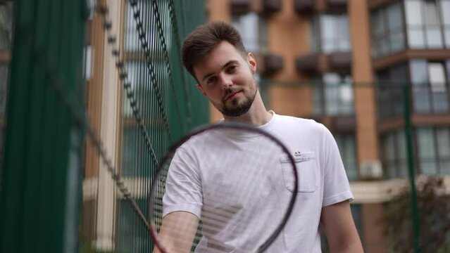 Bottom view zoom in portrait of smiling confident sportsman posing with tennis racket in slow motion. Happy fit Caucasian young man looking at camera standing on outdoor court in urban city