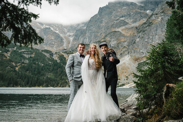 A friend of the wedding couple standing on the stony shore. The bride and groom near the lake in the mountains. Newlyweds together against the backdrop of a mountain landscape. © Serhii