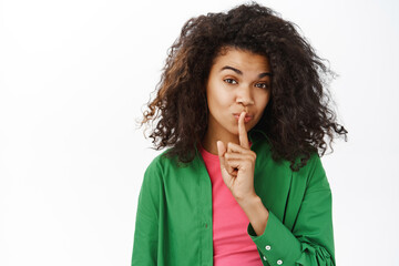 Fototapeta na wymiar Shh keep it secret. Young teenage curly girl, makes hush, shush taboo sign, press one finger to lips and looks mysterious, white background