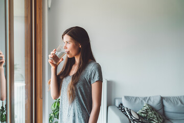 Young woman with long brown hair standing by the window looking out while drinking a glass of water and taking her vitamin pills  - Powered by Adobe