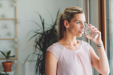 Portrait of blonde woman with a pony tail drinking a glass of water while standing by the window of...
