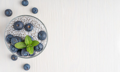 Top view of dessert made of organic chia seeds soaked in plant based milk with topping of ripe blueberries and fresh mint leaf served in glass on white wooden background with copy space for breakfast