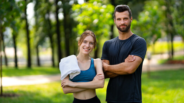 Outdoors fitness training. Portrait image of smiling young couple, woman with man or male bearded coach trainer, standing with folded hands in public park. Sport, cross fit, healthy lifestyle concept.