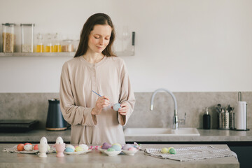 Young woman decorating Easter eggs using brush and paint on the kitchen.