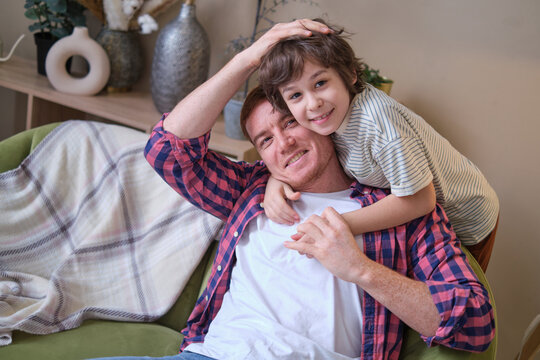 Cheerful father and son moment: wrapped in a warm hug on a cozy couch, beaming at the camera. Navigating the Joys and Challenges of Fatherhood.