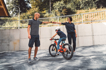 Family of three having fun outside, girl riding a bike, while parents hold hands for their daughter to ride underneath 