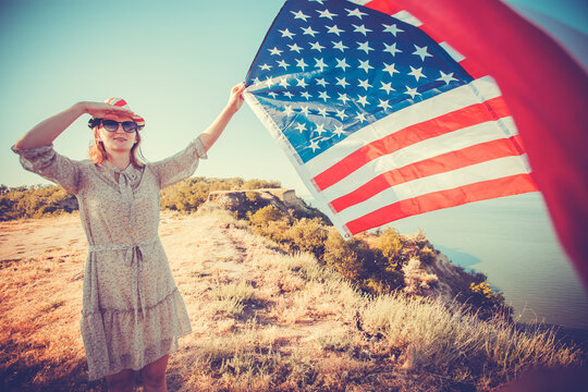 Beautiful young girl holding an American flag on the wind in a meadow. Summer landscape against the blue sky.