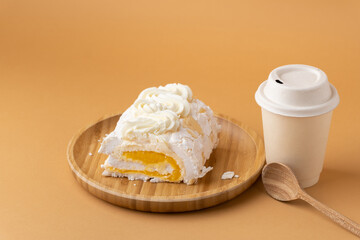 Meringue roll filled with mango puree on the brown background. Meringue roulade with yellow fruit filling in the bamboo wooden plate next to beige paper cup of coffee or tea. Sweet pastry concept